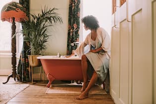 Clean Beauty: Gepflegt auf Nummer sicher gehen A beautiful woman perches on the side of an elegant red roll top bathtub, and waits for it to fill up. She wears a silky robe. Light floods through the window, backlighting the relaxing scene and giving it a dreamy vibe.
