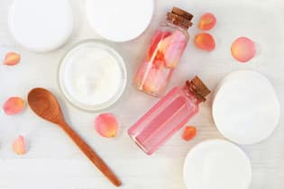 So bringt Rosenwasser Glanz in unsere Haare Tonic and cream, cotton pads, bottles, fresh pink flowers, wooden table preparation background.