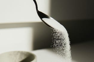 7 Tricks für weniger Zucker im Alltag Zucker fliesst aus einem Löffel in ein Tongefäss