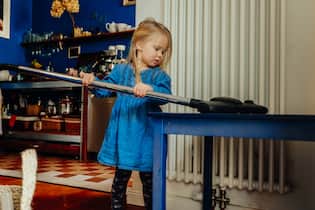 Ab wann kann ein Kind im Haushalt mithelfen? Kid cleaning with vacuum cleaner a table at home.
