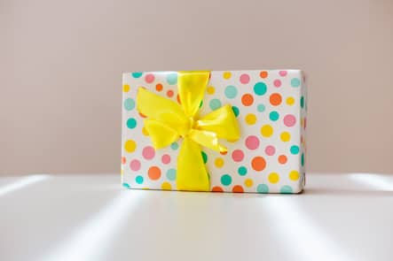 White gift box in a colored pattern with a yellow bow on white desk.