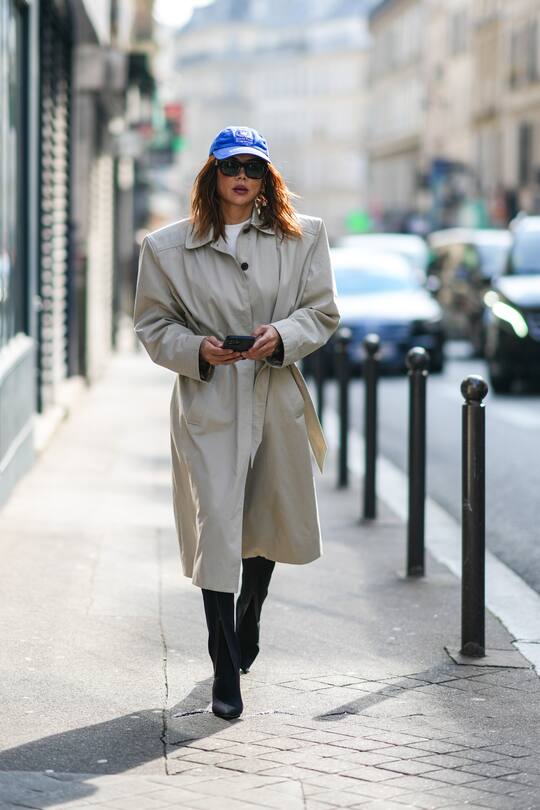 PARIS, FRANCE - MARCH 05: Christine Centenera wears a blue with white embroidered pattern cap from Balenciaga, black sunglasses, a white t-shirt, a beige long / belted with shoulder-pads coat, black shiny leather pointed / heels knees boots / high boots, a gold ring, outside Vivienne Westwood , during Paris Fashion Week - Womenswear F/W 2022-2023, on March 05, 2022 in Paris, France. (Photo by Edward Berthelot/Getty Images)