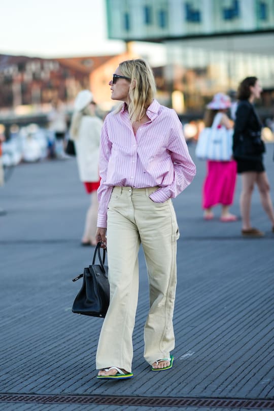 COPENHAGEN, DENMARK - AUGUST 11: A guest wears black sunglasses, earrings, a gold necklace, a pale pink with black and white striped print pattern shirt, beige wide legs denim jeans pants, a black shiny leather large Birkin handbag from Hermes, green / blue / white / yellow plastic flip flop , outside Ganni, during Copenhagen Fashion Week Spring/Summer 2023, on August 11, 2022 in Copenhagen, Denmark. (Photo by Edward Berthelot/Getty Images)