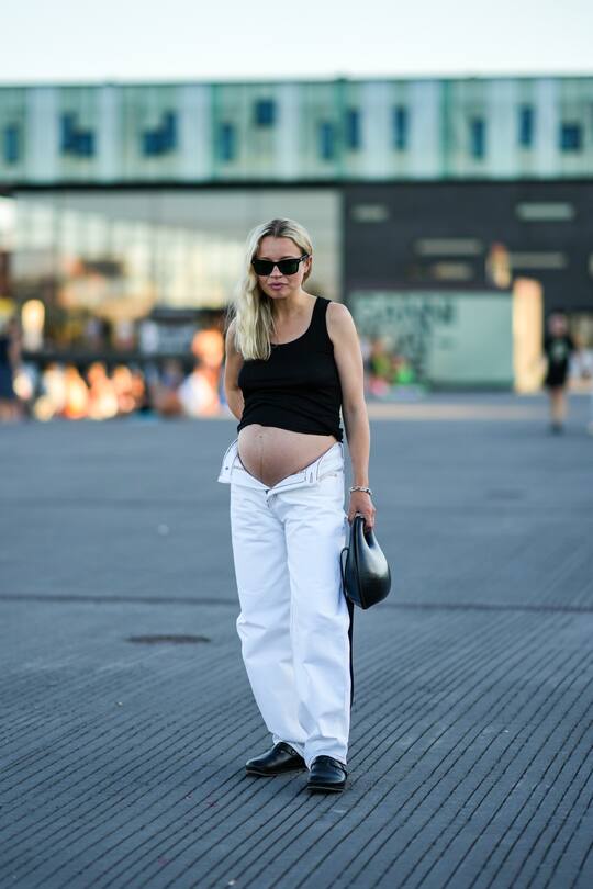 COPENHAGEN, DENMARK - AUGUST 11: A guest wears black sunglasses, a black tank-top, a silver large chain bracelet, rings, a black shiny leather ball handbag, open white denim large pants, black shiny leather mules / shoes , outside Ganni, during Copenhagen Fashion Week Spring/Summer 2023, on August 11, 2022 in Copenhagen, Denmark. (Photo by Edward Berthelot/Getty Images)