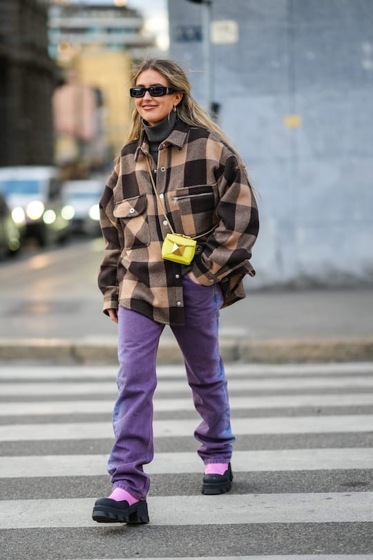 MILAN, ITALY - FEBRUARY 25: Emili Sindlev wears black sunglasses, gold flower pendant earrings, a dark gray ribbed turtleneck pullover, a beige and dark brown checkered print pattern oversized shirt coat, a yellow shiny leather with a large gold stud crossbody bag, purple and blue denim faded large jeans pants, black and pale pink leather block heels ankle boots, during the Milan Fashion Week Fall/Winter 2022/2023 on February 25, 2022 in Milan, Italy. (Photo by Edward Berthelot/Getty Images)