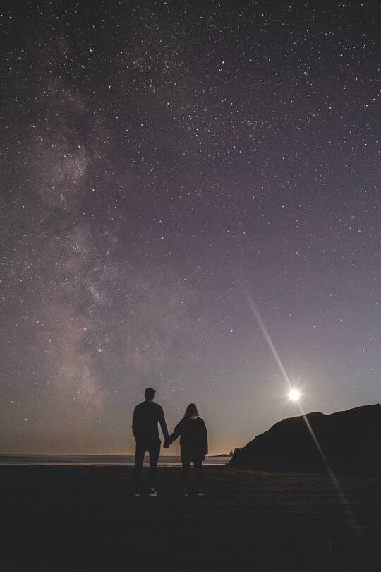 A couple holding hands on a beach with the Milky Way and moon shining in the background