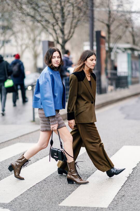 PARIS, FRANCE - MARCH 01: Madelynn Furlong (L) wears a turtleneck brown and blue tweed dress, a lavender-blue leather jacket, a black Prada bag, brown pointy lace-up boots ; Erika Boldrin (R) wears a golden necklace, a brown pantsuit with black velvet cuffs, black velvet insert at the shoulder, wide-leg pants, black pointy boots,outside Nina Ricci, during Paris Fashion Week Womenswear Fall/Winter 2019/2020, on March 01, 2019 in Paris, France. (Photo by Edward Berthelot/Getty Images)