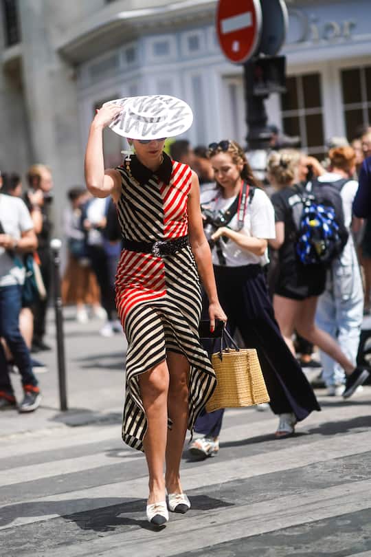 PARIS, FRANCE - JULY 01: A guest wears a straw hat, sunglasses, large earrings, a red, black and beige striped asymmetric dress, a black woven belt, a straw basket, black and beige pointy heeled mules, outside Dior, during Paris Fashion Week -Haute Couture Fall/Winter 2019/2020, on July 01, 2019 in Paris, France. (Photo by Edward Berthelot/Getty Images)