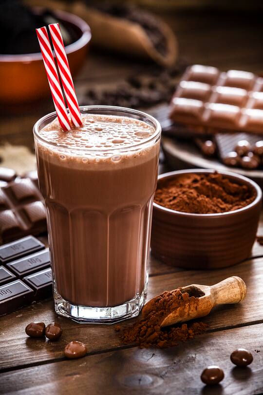 Preparing homemade chocolate milkshake. Chocolate milkshake glass shot on wooden kitchen table. The milkshake glass has two red and white drinking straws inside. A brown bowl filled with ground cocoa is beside the glass. The milkshake glass is surrounded by several chocolate bars. Predominant color is brown. Low key DSRL studio photo taken with Canon EOS 5D Mk II and Canon EF 70-200mm f/2.8L IS II USM Telephoto Zoom Lens