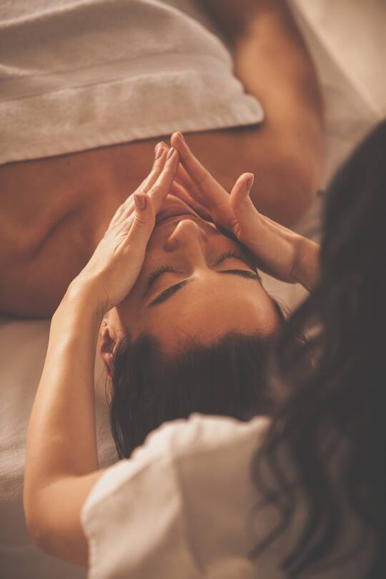 Caucasian woman is lying on massage table and receiving facial massage by a spa therapist.