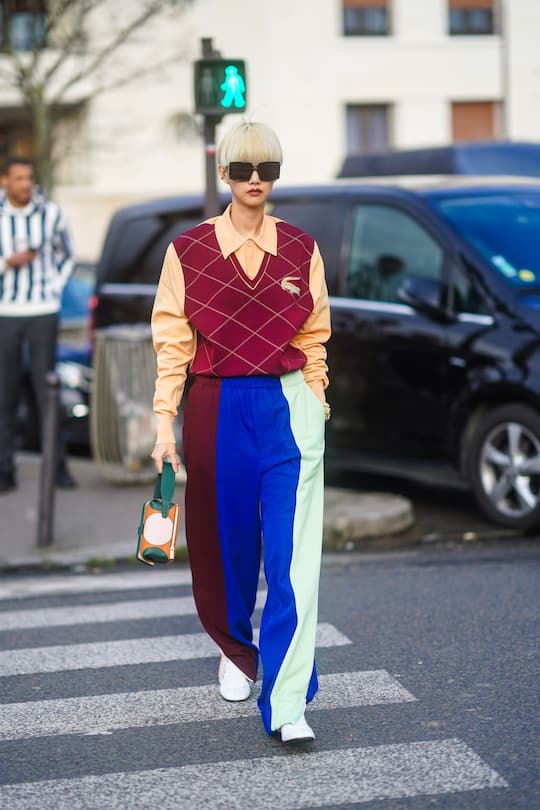 PARIS, FRANCE - MARCH 03: A guest wears sunglasses, a pale orange polo shirt, a sleeveless v-neck Lacoste argyle print burgundy pullover, blue green purple striped pants, a bag with green handle, white sneakers, outside Lacoste, during Paris Fashion Week - Womenswear Fall/Winter 2020/2021 on March 03, 2020 in Paris, France. (Photo by Edward Berthelot/Getty Images)