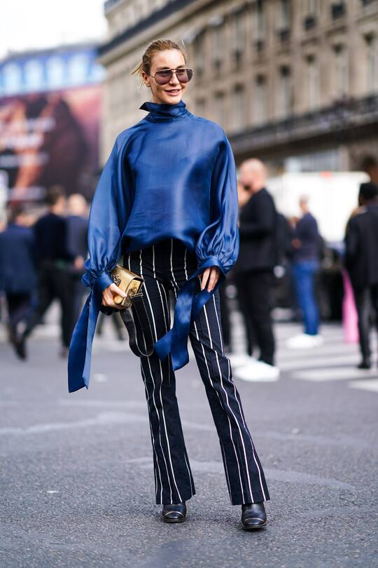 PARIS, FRANCE - SEPTEMBER 27: A guest wears sunglasses, a blue hi-neck lustrous top with long sleeves, black flare pants with white stripes, black boots, a shiny golden handbag, outside Balmain, during Paris Fashion Week - Womenswear Spring Summer 2020 on September 27, 2019 in Paris, France. (Photo by Edward Berthelot/Getty Images)