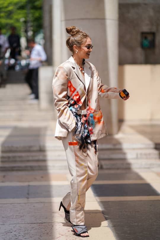 PARIS, FRANCE - JUNE 18: Gigi Hadid wears sunglasses, earrings, necklaces, a Heron Preston lustrous beige pantsuit with colorful paintings print, grey padded heeled mules, outside Heron Preston, during Paris Fashion Week - Menswear Spring/Summer 2020, on June 18, 2019 in Paris, France. (Photo by Edward Berthelot/Getty Images)