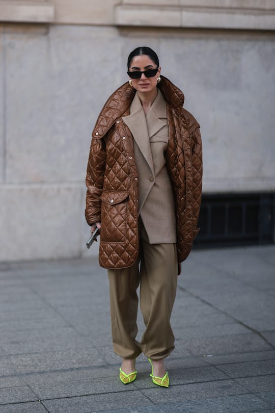 PARIS, FRANCE - MARCH 07: A guest wears black sunglasses, a beige blazer jacket, brown oversized puffy pants, a brown shiny quilted oversized puffer jacket, gold earrings, a khaki shiny leather handbag, neon yellow shiny leather and tulle pointed pumps heels shoes, outside Louis Vuitton, during Paris Fashion Week - Womenswear F/W 2022-2023, on March 07, 2022 in Paris, France. (Photo by Jeremy Moeller/Getty Images)