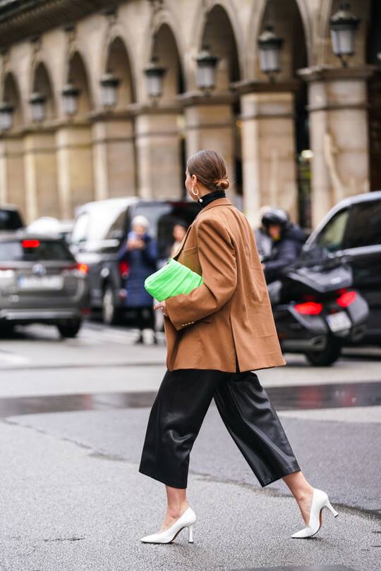 PARIS, FRANCE - MARCH 02: A guest wears earrings, a black turtleneck, a caramel-brown oversized jacket, black leather wide-legs short pants, white pointy heeled pumps, a neon-green clutch, outside Giambattista Valli, during Paris Fashion Week - Womenswear Fall/Winter 2020/2021, on March 02, 2020 in Paris, France. (Photo by Edward Berthelot/Getty Images)