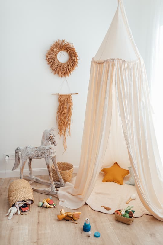 Messy children's playroom full of toys. Beautiful decoration nursery with natural macrame decoration, organic canopy, wicker baskets and wood toys. Vertical photo.