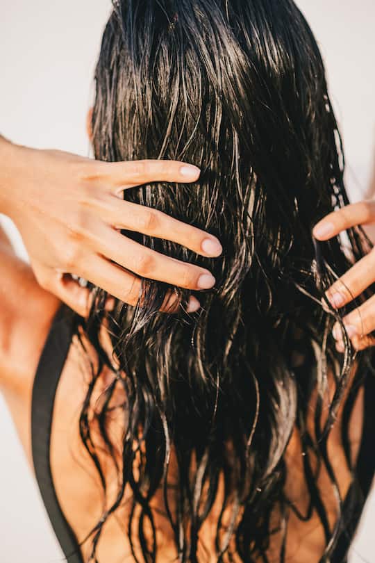 A beautiful, unrecognizable woman washing her hair in an outdoor shower. She is wearing a black swimsuit. Vitamins for healthy hair, skin, and nails. Hydration with natural cosmetics, collagen, and biotin supplements. Normalizing natural manicure, gel-free. Shot from behind