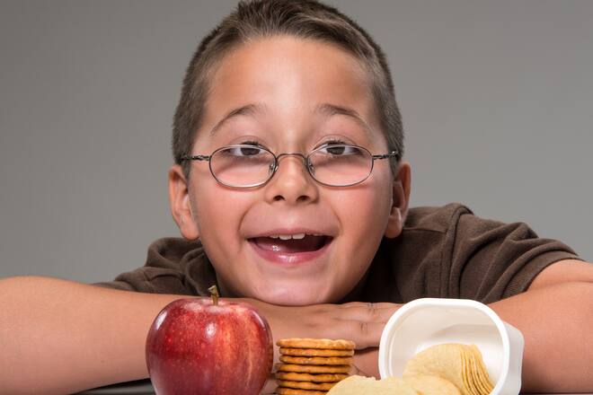 "Schoolboy posing with a red delicious apple, potato chips, and crackers on gray background"
