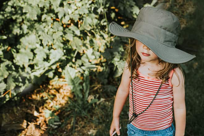 Cute young child wearing a strappy top and a wide brim khaki hat to protect her from the sun. Her face is partly obscured and she is expressionless. Leaves provide a space for copy.