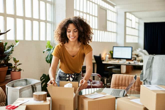 Confident businesswoman packing boxes at table. Young female entrepreneur is working at home office. She is in casuals.