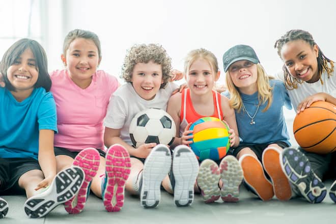 A multi-ethnic group of elementary age children are sitting in a at the gym. They are holding a soccer ball, volleyball, and basketball and smiling while looking at the camera.