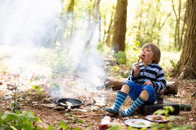 Kind sitzt an Feuer und isst eine Wurst.