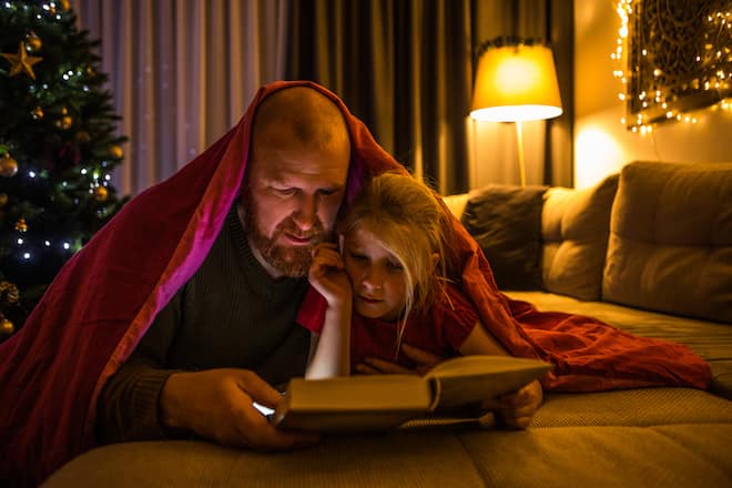 Beautiful blonde daughter and bearded redhead father reading a book together in a family home adorned with christmas decorations