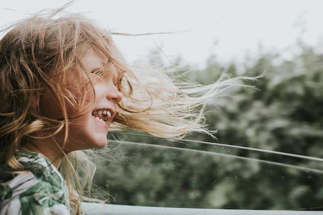 Laughing pre-school age girl enjoys a car journey. She sits beside an open window as the wind rushes through her long hair and it blows around her face. Window provides a space for copy.