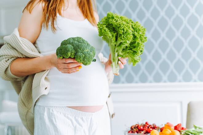 Unrecognizable pregnant woman in white clothes holding a head of broccoli and lettuce in her hands