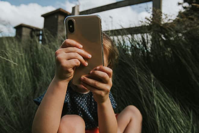 A young child holds a mobile phone up, obscuring her face, and takes a photo of the photographer, or takes a selfie. The scene is outside, with grass providing space for copy.