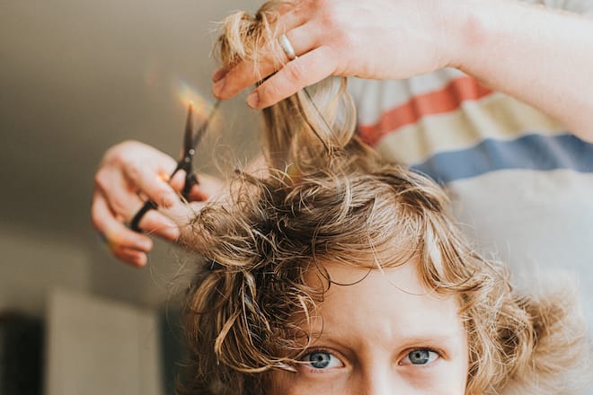 Comical image of young boy having a lockdown haircut at home, during the Covid Pandemic. Boy looks directly at the camera showing some uncertainty at the situation, while father is unqualified to cut the child's long, blond curly hair. Space for copy.