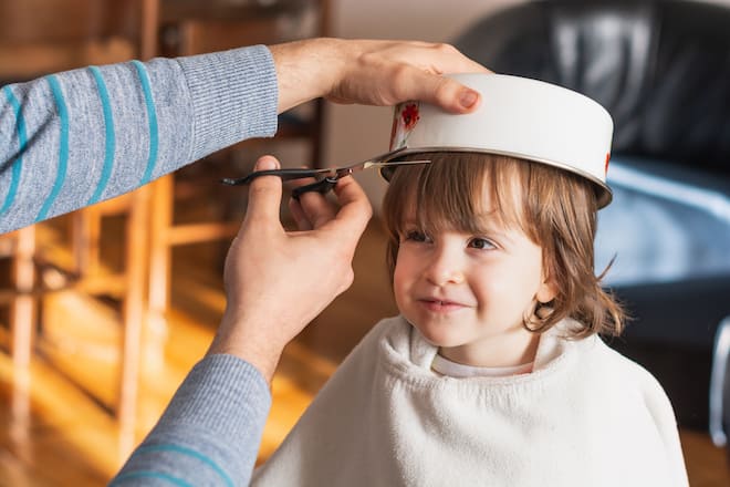 Smiling little girl getting haircut from her father wearing pot on her had