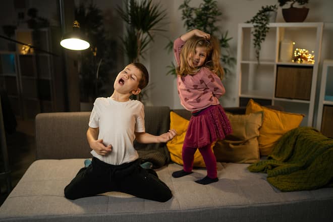 Playful brother and sister dancing and singing in living room on sofa
