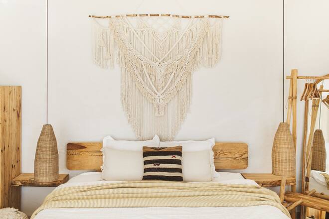 Cozy interior of the house is done in white and beige colors: craft wicker lamps, handicrafted macrame panno hanging on the wall and bed with cozy soft fluffy pillows and blanket. Bedroom decoration in boho style. Hygge style. Morning mood.
