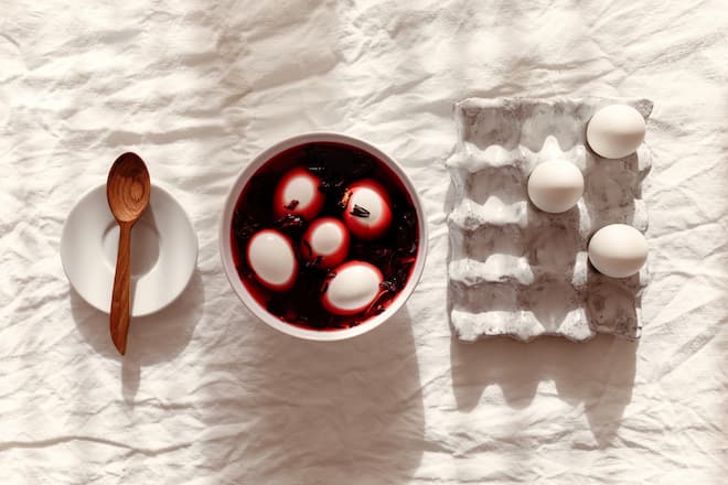 Woman's hands dyes eggs with organic dye from Hibiscus herbal tea use as an infusion from crimson or deep magenta-colored. Top view of Easter eggs being colored in bowl with natural Hibiscus tea leaves dye. Easter tradition.