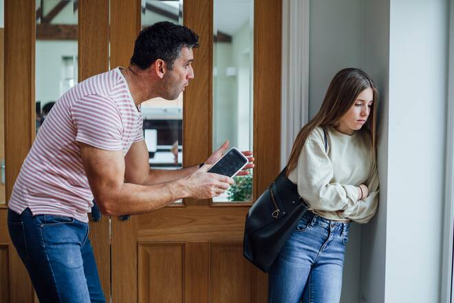 A father and his teenage daughter discussing her behaviors in the passageway of the family home he has confiscated her phone because her bill was so high.