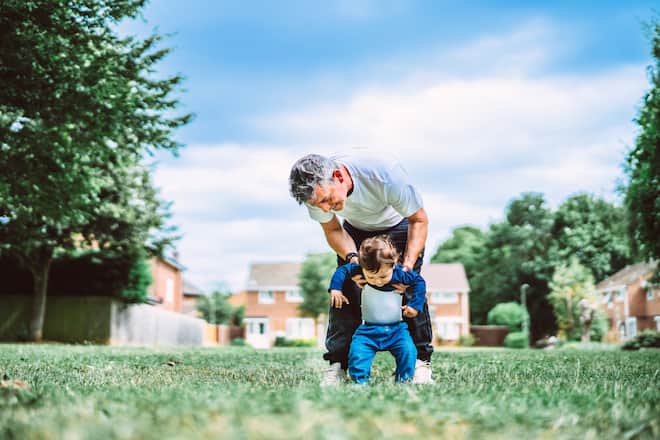 Father carefully guiding his toddler son as they take their first steps on the grass. This photograph is perfect for illustrating themes of early childhood, parent-child bonding, support, and the milestones of growing up.