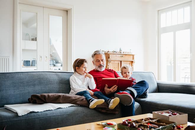 Two young grandchildren sitting on a large sofa and reading a festive book with their grandpa, getting ready for Christmas.