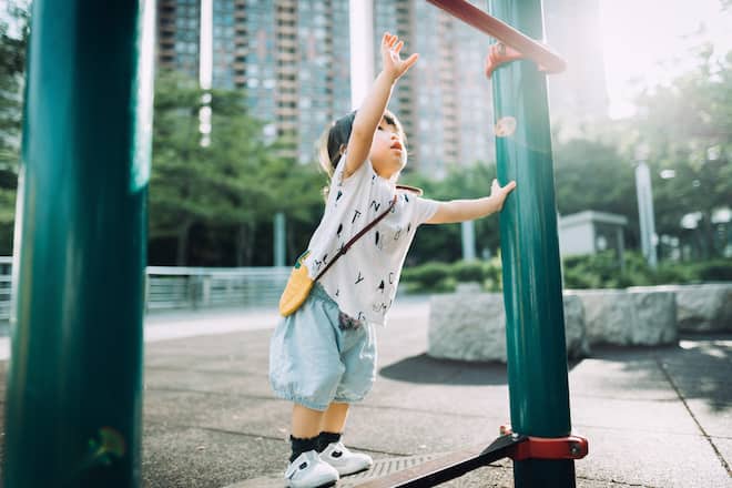 Playful little toddler girl arms raised and trying to reach a pull up bar at the outdoor playground on a lovely sunny day