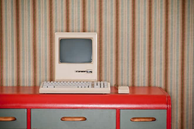 Old classic computer sitting on an art deco retro desk. The wall is covered in a wallpaper with a striped wallpaper.*