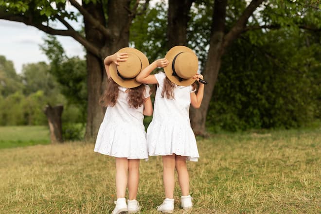 Back view of twin sisters in similar white dresses touching straw hats and admiring tall trees on summer weekend day in park
