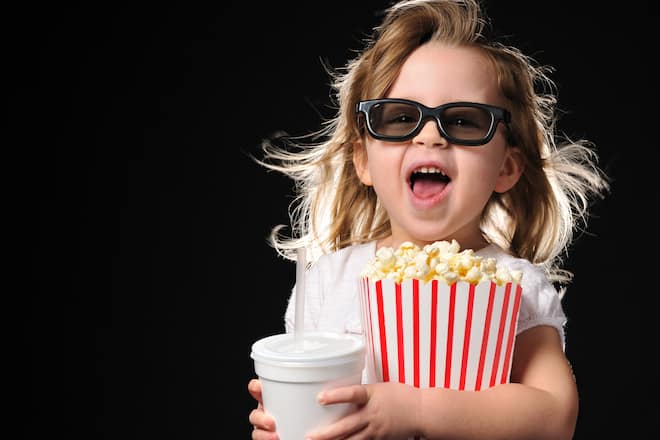 Horizontal studio image of a cute young girl wearing 3D glasses and holding on tightly to her theater popcorn and soda. She is obviously very excited to be watching the 3D technology. Her mouth is open as if she's squealing with enjoyment.
