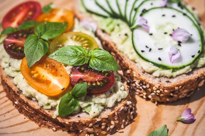A healthy breakfast or snack, slices of whole grain bread smeared with avocado and slices of cucumber and cherry tomatoes, topped with edible flowers, nigella seeds and fresh basil.