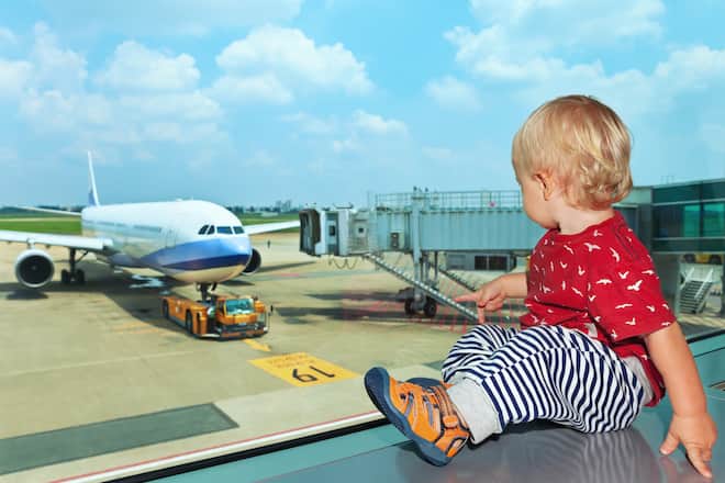 Little baby boy waiting boarding to flight in airport transit hall and looking through the window at airplane near departure gate. Active family lifestyle, travel by air with child on summer vacation