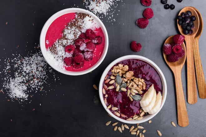 Breakfast smoothie bowls with frozen blueberries and raspberries, topped with bananas, nuts, sunflower seeds and coconut flakes, top view
