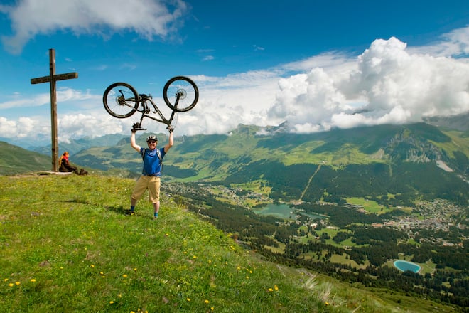 A mountain biker is standing on a summit with his bike in the air. Mountainbiking in Lenzerheide. The region of Graubunden in Switzerland is a single track paradise.