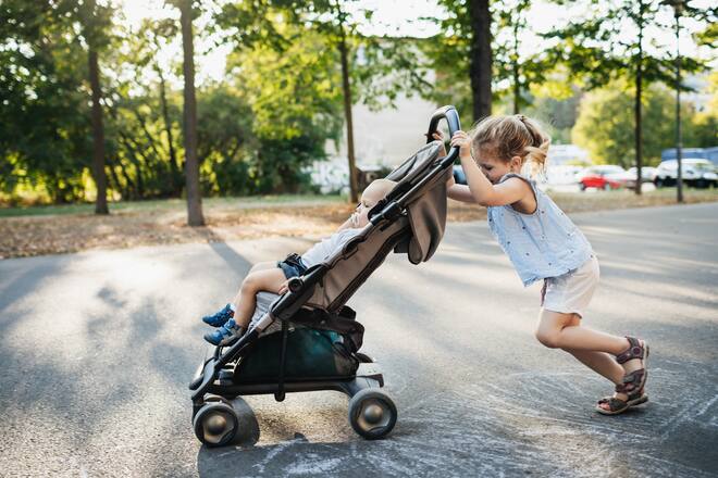 A young girl pushing her baby brother in a stroller while on a day out in the park together.