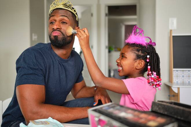 An African American father and daughter playing dress up and putting on makeup n their home.