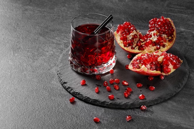 A transparent glass full of sweet pomegranate juice and a cut garnet on a round plate on a dark gray background. Summer, healthful and fruity red cocktail with black straw and fresh garnet.