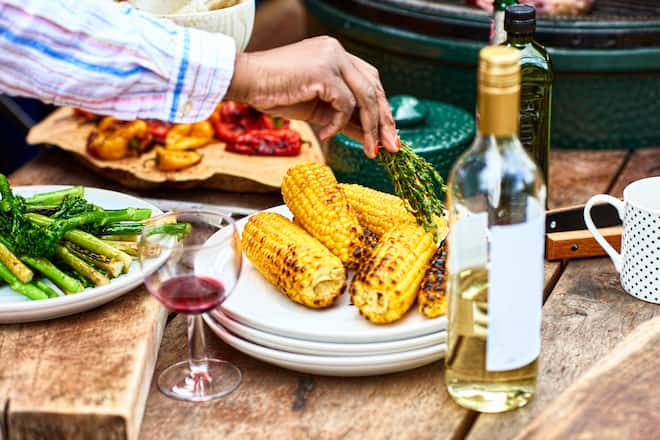 Char grilled corn with green vegetables and wine on table, woman seasoning food with herbs, food preparation, healthy eating
